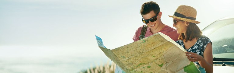 A married couple looking at a large map for their next road trip destination as they stand in front of their car parked on the side of a beachside road