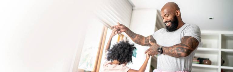 A stepdad holds his daughter's hands as they dance together around the living room of their blended-family home