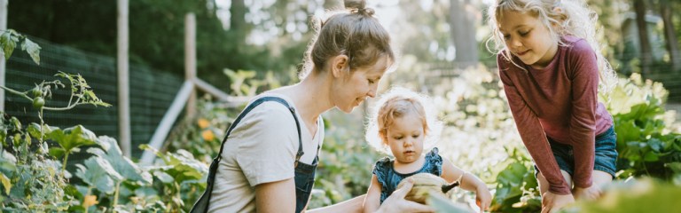 A mom is with her two young girls in their garden looking at a squash she is holding and at the same time is helping them understand how to deal with anger issues