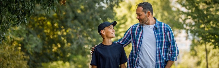 A father with his arm around his son while they're walking under some shade trees and having a father-son talk about life issues